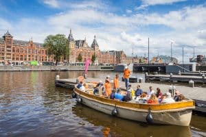 People enjoy a group boat tour on an Amsterdam canal in front of a historic red-brick building on a sunny day.