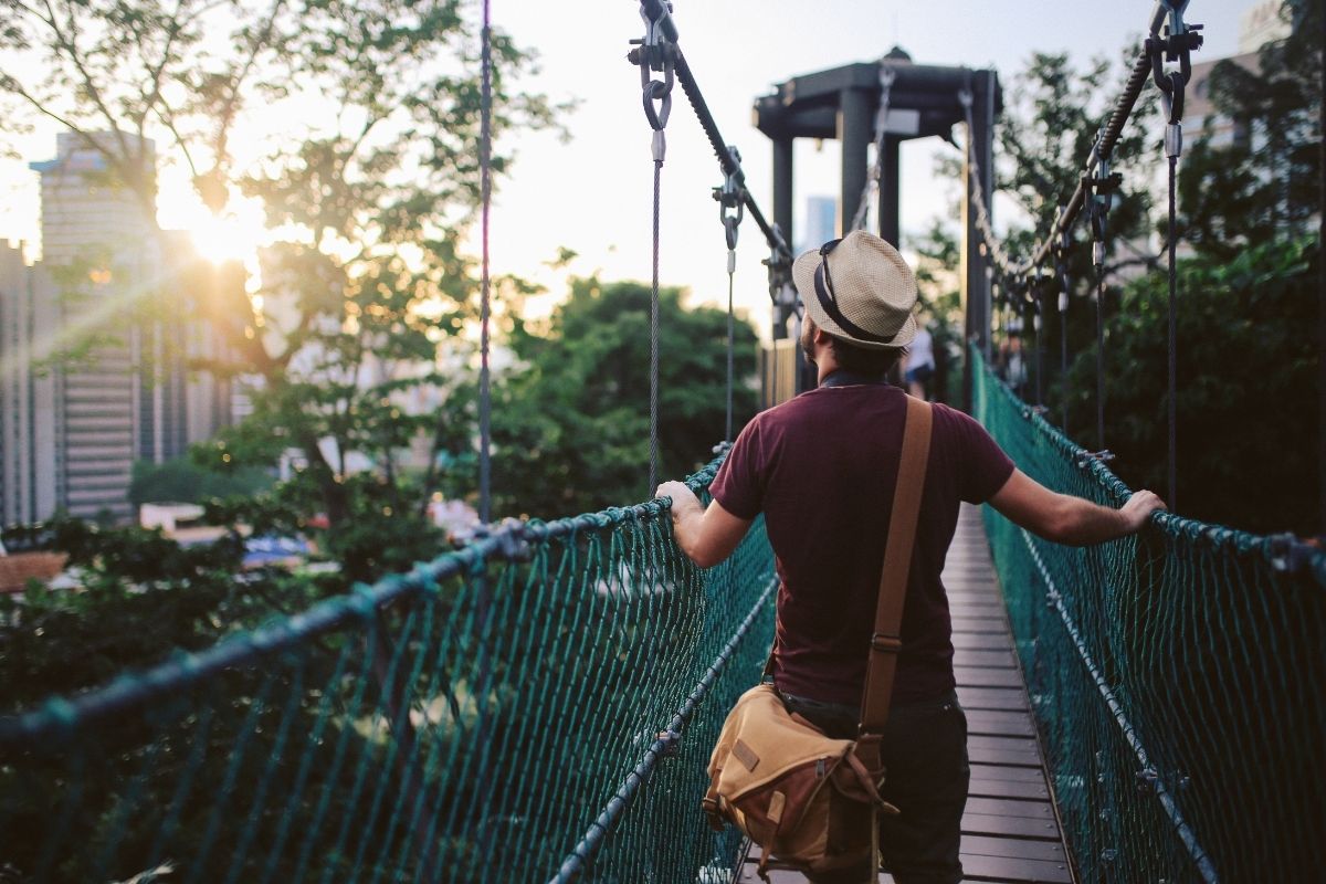 Person in a hat walking on a suspension bridge with trees and city skyline in the background at sunset.